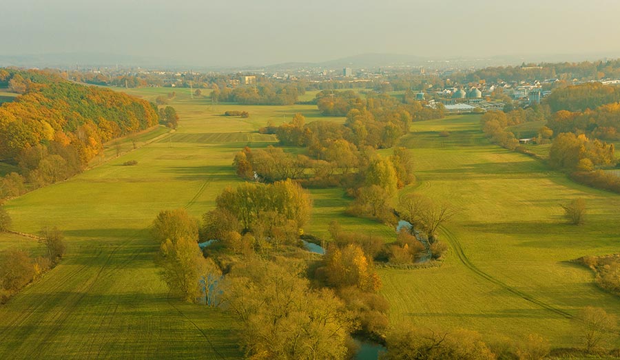 Herbstliche Flusslandschaft mit Wiesen, Bäumen und Stadt im Hintergrund aus der Vogelperspektive