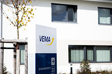 VEMA Firmenschild vor modernem Bürogebäude mit großen Fenstern und kleinem Baum